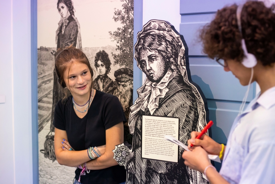Wystawa stała w Muzeum POLIN. Dziewczyna patrzy na chłopaka, który coś notuje. Oboje są w słuchawkach.