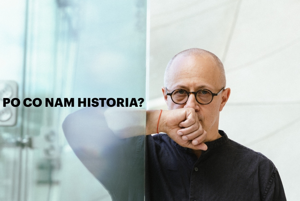 Mikołaj Grynberg oparty o szklaną ścianę w holu głównym Muzeum POLIN. Trzyma dłoń na brodzie.