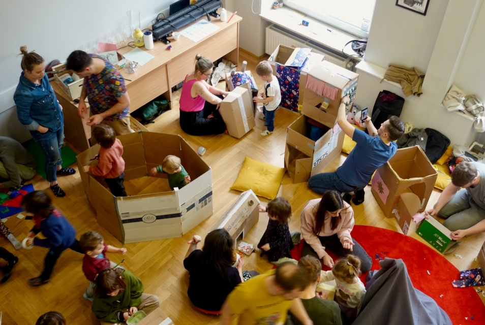 Dzieci i dorośli na zajęciach w klubie Kinderwelt. Niektóre dzieci siedzą w kartonowych domkach.