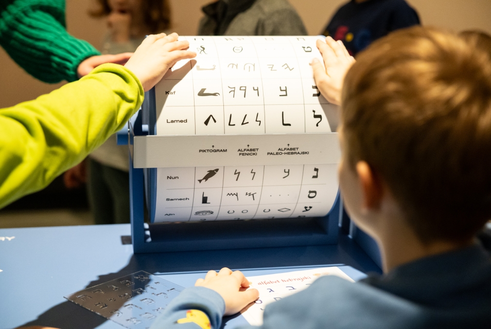 Chłopiec obraca wałek edukacyjny z nazwami słów i literami w różnych językach żydowskich.