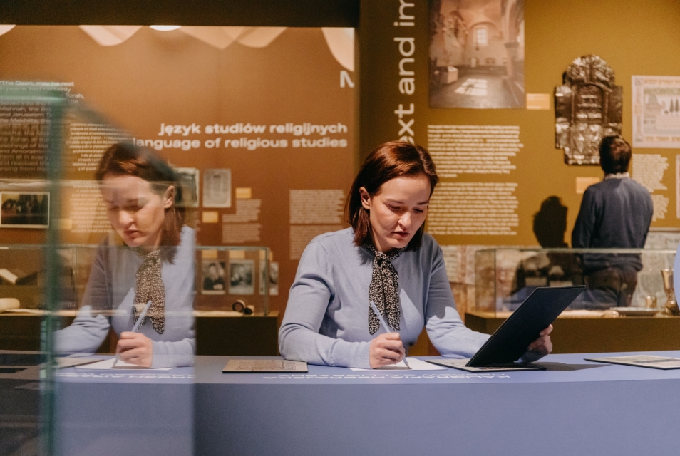 Wystawa "Moc słów. O językach żydowskich". Kobieta siedzi przy stole i pisze coś na kartce. W drugiej dłoni trzyma tabliczkę, z której przepisuje tekst. Za nią stoi mężczyzna zwiedzający wystawę.