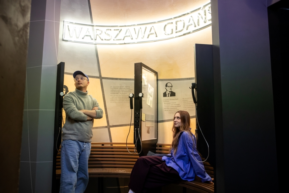 Mężczyzna stoi z założonymi rękami, a kobieta siedzi na ławce na wystawie stałej w Muzeum POLIN.