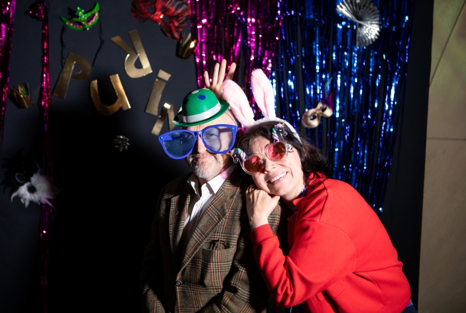 Kolorowa ścianka z napisem Purim. Na jej tle mężczyzna w dużych niebieskich okularach i z zielonym kapeluszem na głowie. O jego ramię opiera się kobieta w różowych okularach w jednorożce i uszami zająca na głowie.