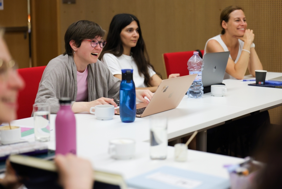 A group of women are sitting at a table in a conference room. They are holding laptops with open screens in front of them.