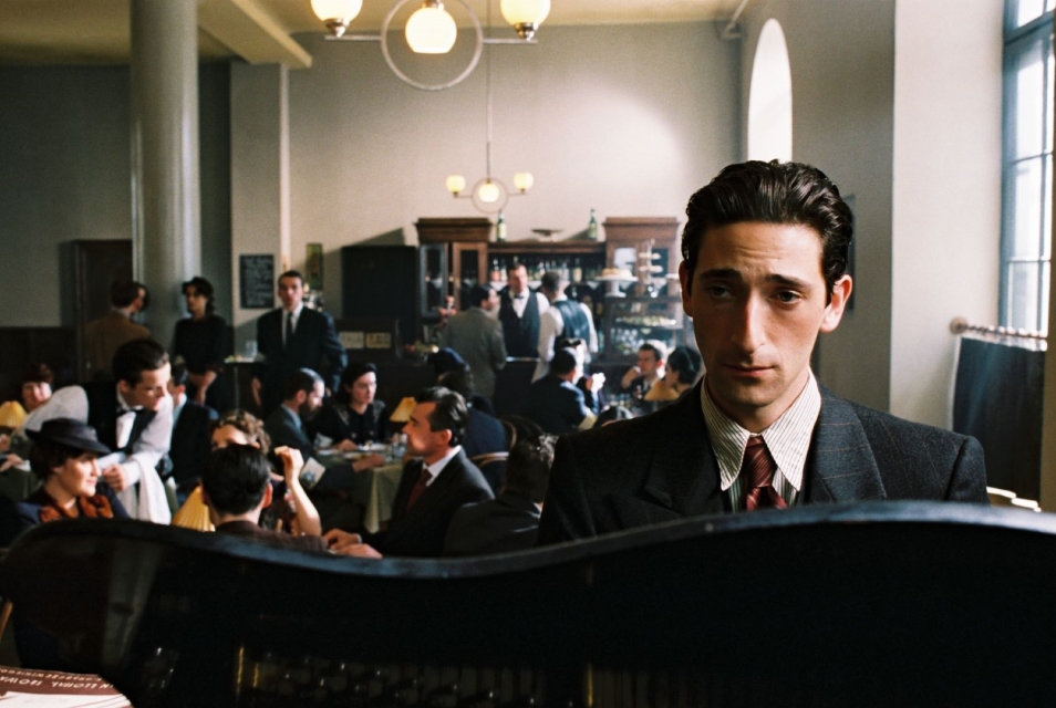 Kadr z filmu "Pianista". Tytułowy bohater siedzi przy fortepianie w kawiarni pełnej gości.