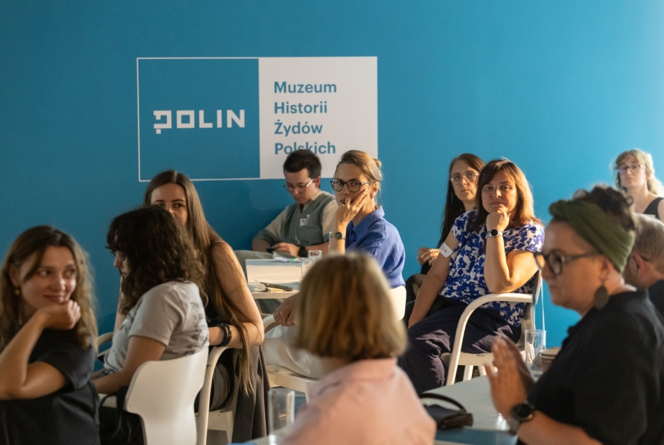 Uczestniczki i uczestnicy muzealnego think-tanku siedzą przy stolikach. W tle logo Muzeum POLIN.
