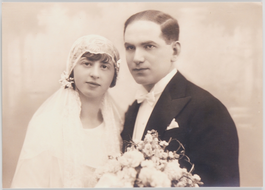 Fotografia ślubna Ernestyny z domu Kutin i Izydora Marka Bauera. Kobieta jest ubrana w suknię z dekoltem w kształcie litery V. Na głowie ma czepiec ślubny z welonu z koronki ozdobiony koralikami. Mężczyzna ma na sobie marynarkę, kamizelkę i koszulę, pod szyją ma muszkę.
