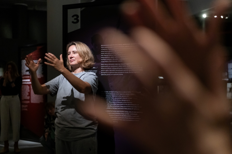 In the foreground, blurred hands. In the background, a woman with her hands raised.