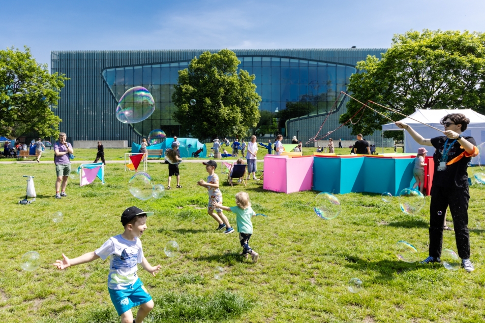 A group of children in the square in front of the POLIN Museum on Children’s Day. Soap bubbles are floating around them, blown by an adult woman.