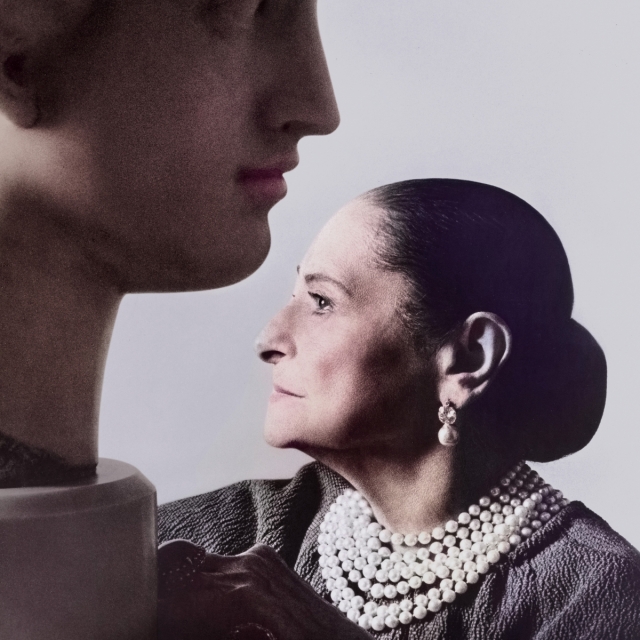 A profile portrait of Helena Rubinstein next to a large bust of a man. The woman has her hair smoothly combed back into a bun, and she is wearing a pearl necklace and earrings. She is looking straight ahead against a light background.