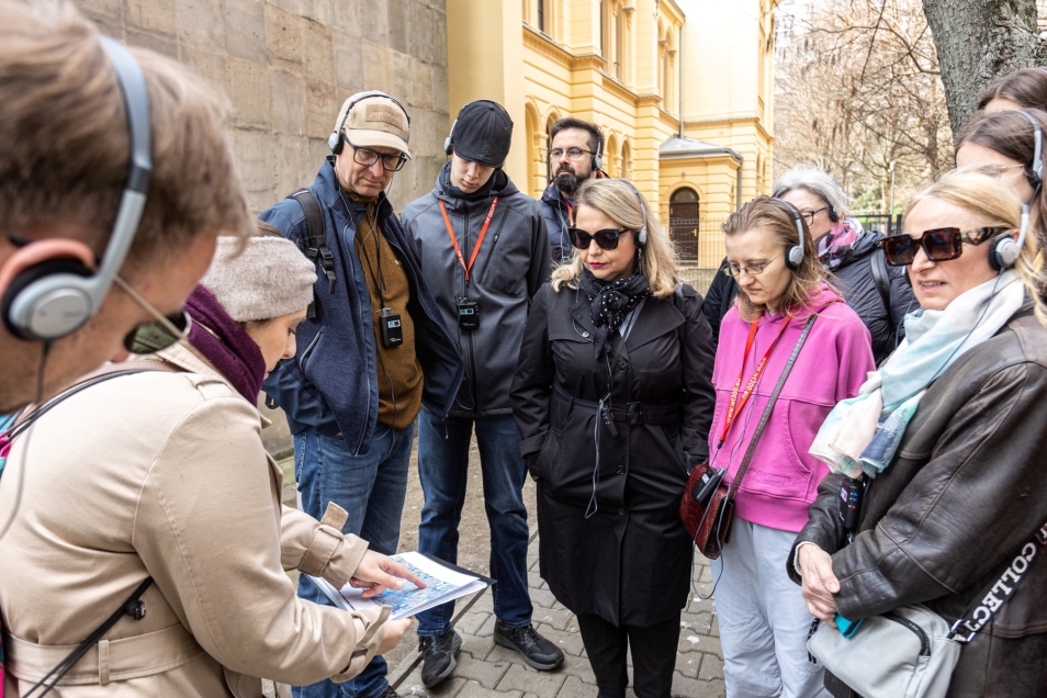 Grupa uczestniczek i uczestników spaceru miejskiego po Warszawie. Przewodniczka pokazuje im coś palcem w swoich notatkach.