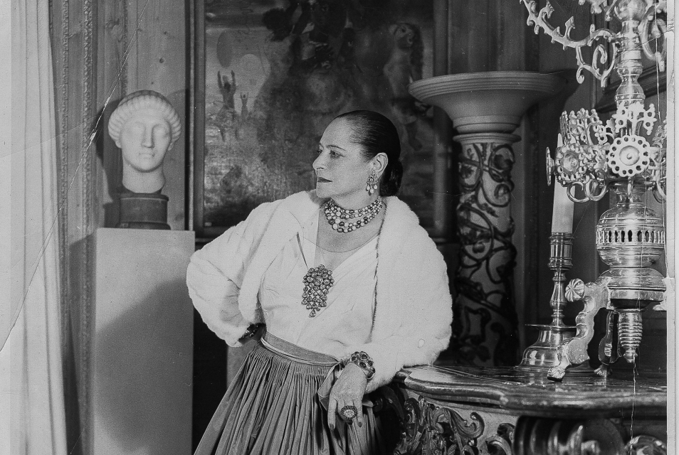 Helena Rubinstein w białej bluzce z bufiastymi rękawami i długiej spódnicy, stoi oparta o ozdobną komodę. W tle dzieła sztuki.