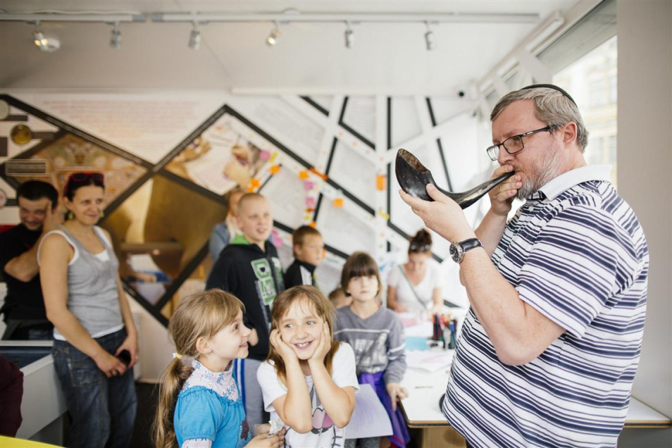 Mężczyzna gra na rogu. Wokół grupa dzieci, jedna dziewczynka zakrywa uszy.