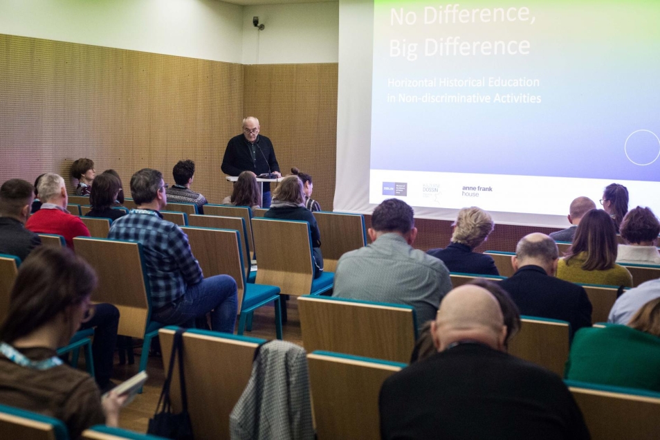 The speech of the Museum POLIN Acting Director Zygmunt Stępiński. Zygmunt Stępiński speaks in the left corner of the room. On the right there is a screen with the white title of the seminar “No Difference, big Difference” on a blue background. In the foreground there are participants of the seminar sitting in the rows.