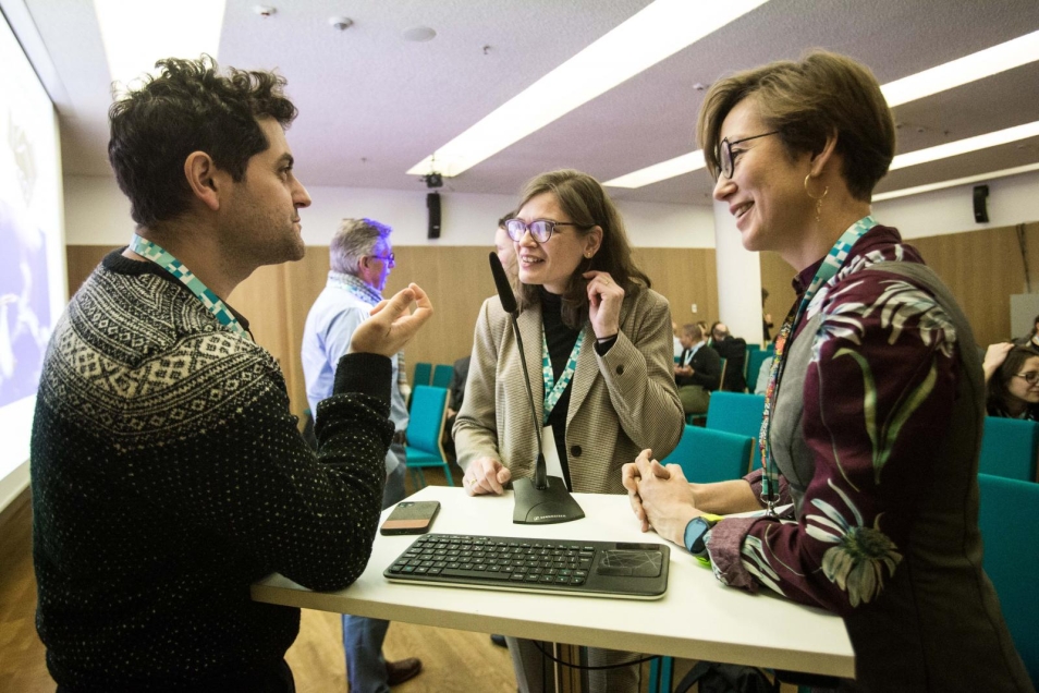 Work in groups during an integration exercise. In the foreground there are three people standing around the table and discussing. On the left side stands a man and on the right side two women. In the background other groups, having a discussion, can be found.