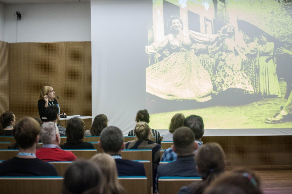 PH.D. Małgorzata Kołaczek’s lecture on romophobia. The picture presents the listeners of the lecture on romophobia. The lecturer stays on the left side of the photo. In the center there is a screen on which a slide showing a photo of the stereotypical representation of Roma women is displayed. Women are dressed in long, patterned skirts, captured while dancing