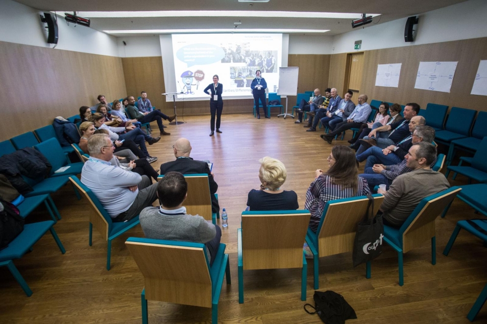 A police group during workshops. The participants of the police workshop are sitting in one large circle in the middle of the room. The facilitators are standing in the middle of the circle. There is a screen behind them. On the screen a slide about the similarities and differences within the police is displayed