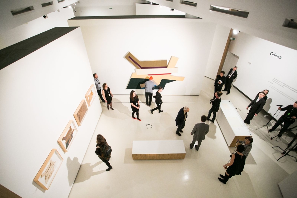 Widok z góry na salę w muzeum Polin. Na ścianach wiszą obrazy. Ogląda je grupa ludzi. Wystawa „Frank Stella i synagogi dawnej Polski”. 