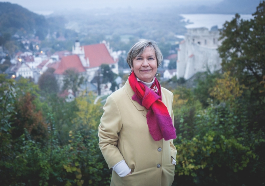 Aleksandra Markiewicz. Siwowłosa kobieta w płaszczu i czerwono-różowym szaliku. Stoi na tle zieleni i zabudowy miejskiej.