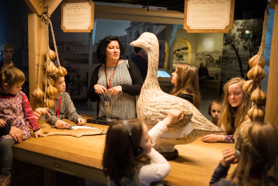 Chanuka w Muzeum POLIN - spacer z przewodnikiem po wystawie dla rodzin z dziećmi - Opis zdjęcia: grupa dzieci stoi wokół drewnianej lady straganu na wystawie muzeum. Słuchają przewodniczki. Na straganie wiszą wieńce czosnku i stoi gęś - wykonane z drewna. Panuje półmrok.