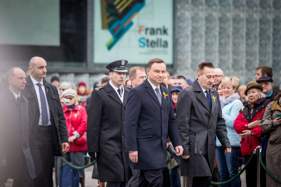 Prezydent Andrzej Duda wraz z kilkoma osobami idą na tle Muzeum Polin. Za nimi osoby uczestniczące w uroczystościach.
