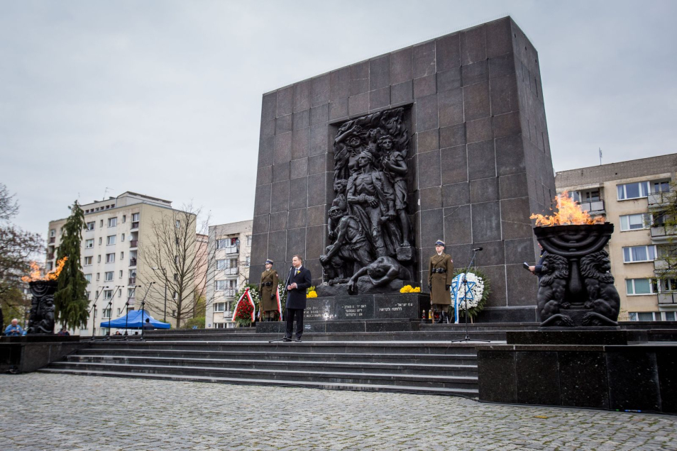 Pomnik Bohaterów Getta w Warszawie z płonącymi pochodniami, wieńcami i żołnierzami stojącymi na warcie. Pośrodku jedna osoba przy mikrofonie.