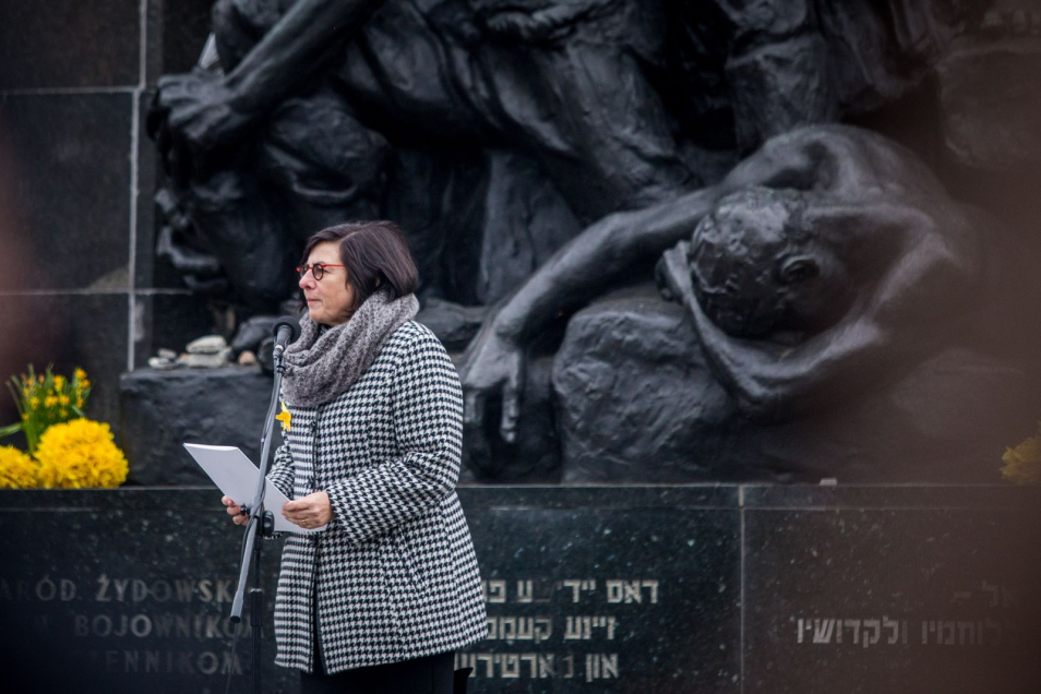 Kobieta stoi przed mikrofonem na tle Pomnika Bohaterów Getta w Warszawie i czyta tekst przemówienia