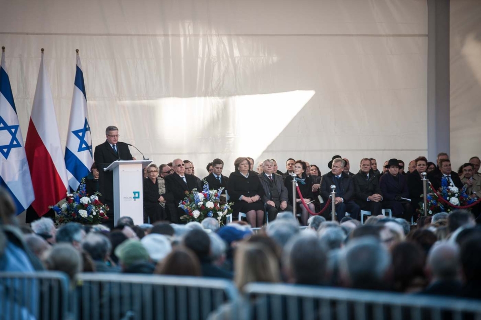 Bronisław Komorowski przy mównicy. Obok siedzą politycy. W tle flagi polskie i izraelskie.