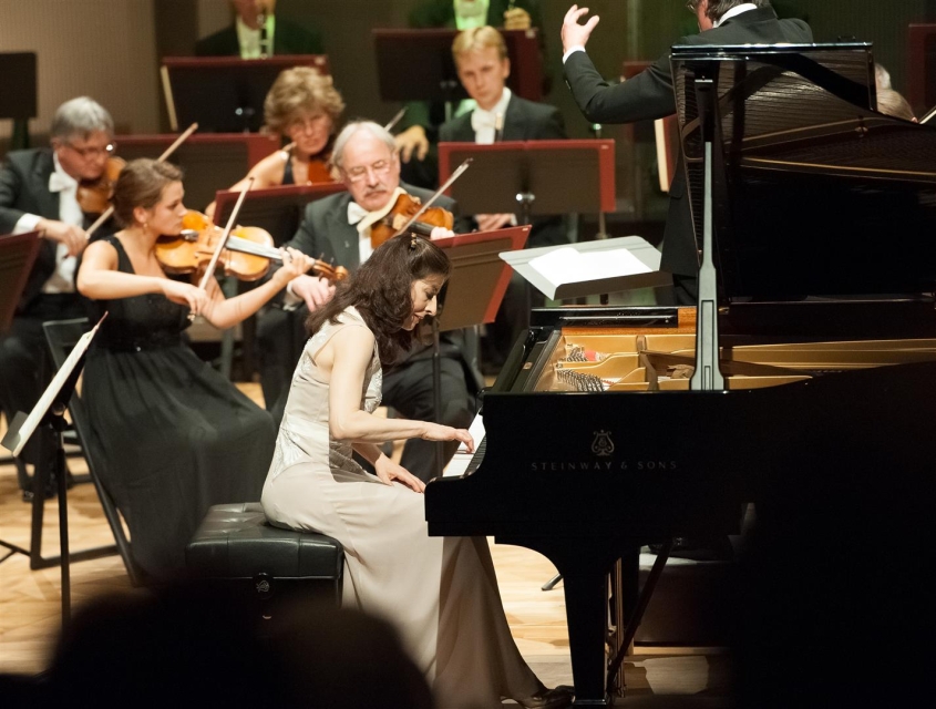 Pianistka gra na fortepianie, który stoi na środku sceny. Pięciu muzyków gra na skrzypcach