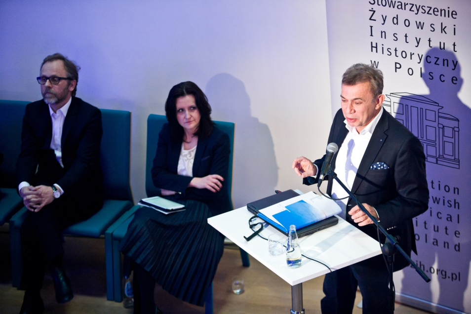 3 osoby - 2 słuchają, jeden z prelegentów przedstawia prezentację.