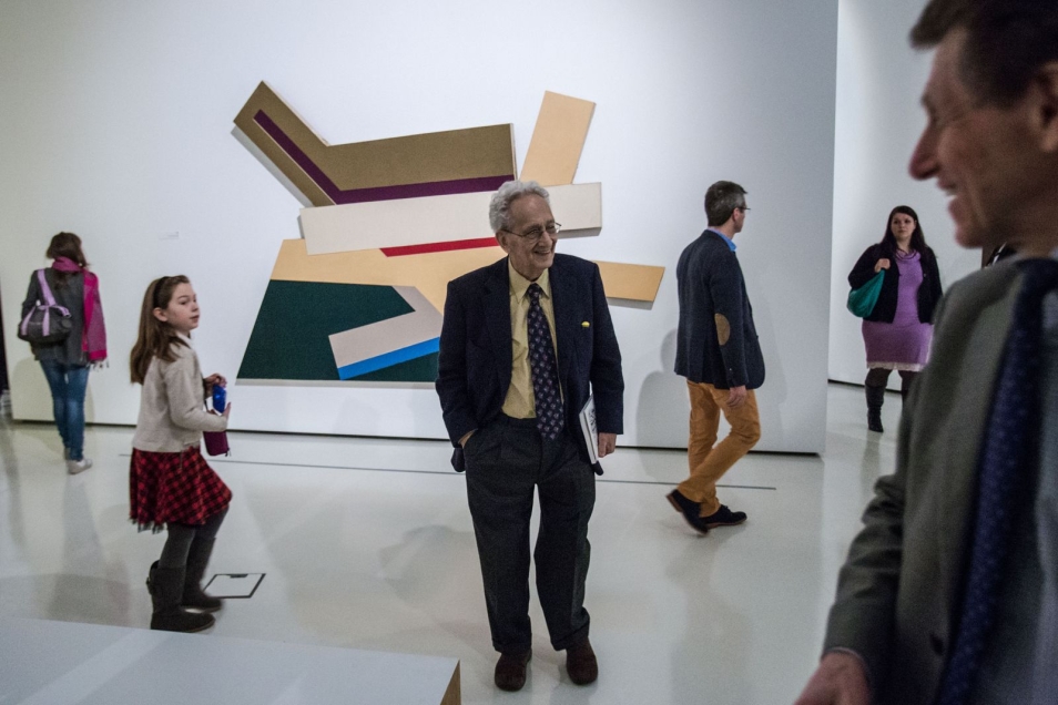Sala w muzeum. Wernisaż wystawy „Frank Stella i synagogi dawnej Polski”. W tle, na ścianie, duży, kolorowy obraz. W sali grupa ludzi.  