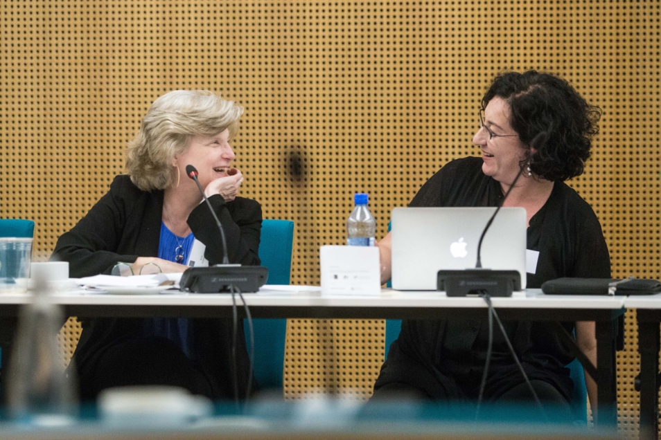 Two women are sitting next to each other at a workbench, facing each other and talking. In front of them are microphones and notes.