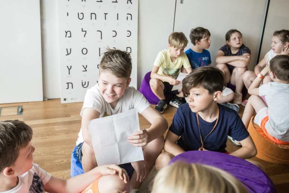 Grupa dzieci w sali, rozmawiają ze sobą. Na pierwszym planie dwóch chłopców - ten po lewej trzyma kartkę i się uśmiecha, drugi po prawej patrzy zasłuchany. Za dziećmi biała plansza z czarnymi hebrajskimi literami. Warsztaty dla szkół podstawowych, klasy 4-6 w Muzeum POLIN