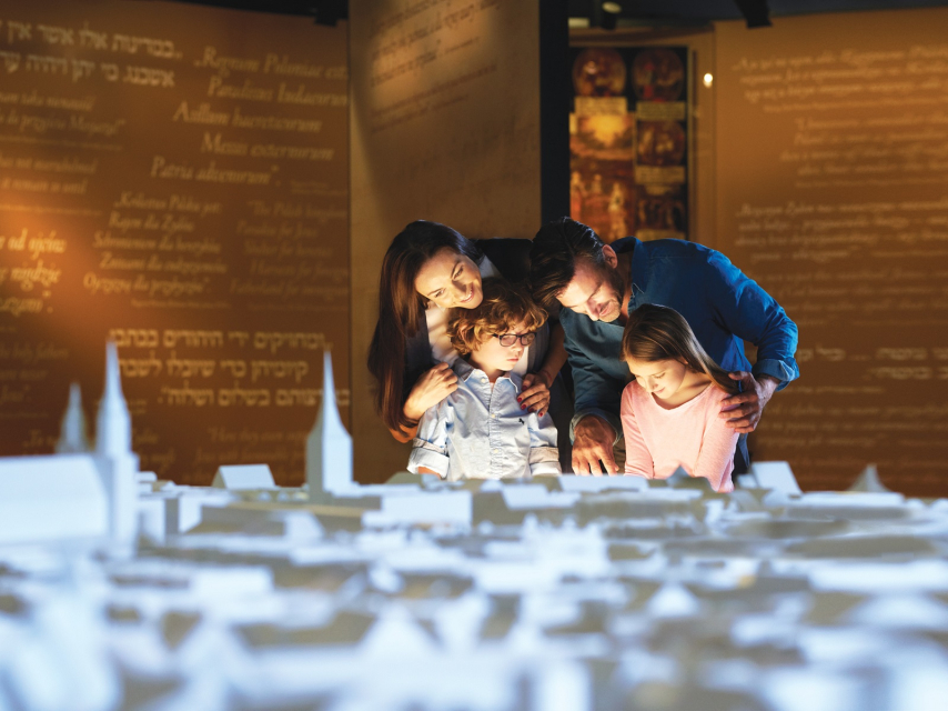Odwiedź wystawę stałą Muzeum POLIN, Na zdjęciu: Kobieta i mężczyzna z dwójką dzieci stoją przed olbrzymia makietą miasta. Makieta jest koloru białego i znajduje się w przestrzeni wystawy stałej Muzeum POLIN