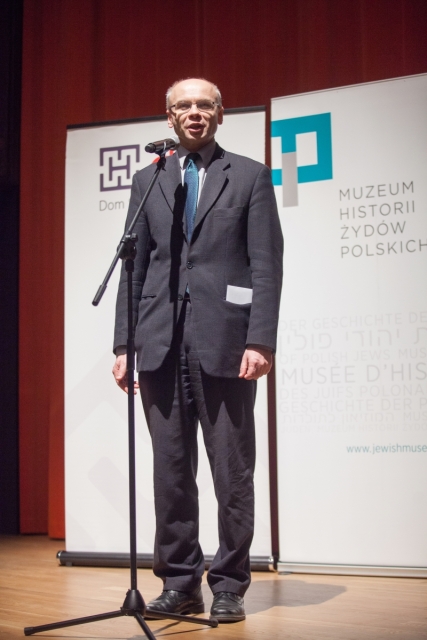 Dariusz Stola stoi przy mikrofonie, w tle plansza z logo Muzeum POLIN i Domu Spotkań z Historią.