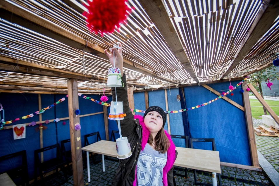 Sukkot - spacer z przewodnikiem po wystawie stałej Muzeum POLIN dla rodzin z dziećmi - Dziewczyna stoi w przygotowanym na Sukkot namiocie - ściany niebieskie, dach z mat trzcinowych. Wiesza pod dachem ozdoby świąteczne, w postaci ozdobionych dzwonków. Ubrana jest w bluzę i czapkę - jesiennie