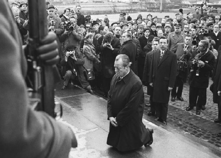 Zdjęcie archiwalne. Willy Brandt klęczy, w tle grupa ludzi.