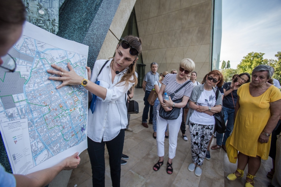 Wędrujący Uniwersytet Muranowski - kobieta pokazuje innym osobom coś na mapie.
