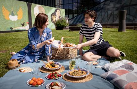 Kobieta i mężczyzna piknikują na łące przed Muzeum POLIN. Na trawie leży koc, na którym znajdują się miski z jedzeniem.