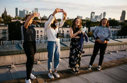 Na dachu budynku stoją rzędem cztery osoby w letnich ubraniach. Każda trzyma przed sobą telefon lub aparat fotograficzny. Robią zdjęcia. W dole za nimi panorama miasta.