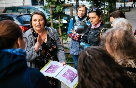 Agnieszka Kuś trzyma w ręku graiki z planem Muranowa i opowiada zawartą w nich historię zgromadzonym wokół niej uczestnikom spaceru