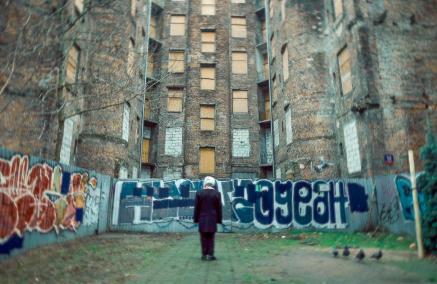 Starsza kobieta stoi przed starą zniszczoną kamienicą, ogrodzoną blachą z graffiti.