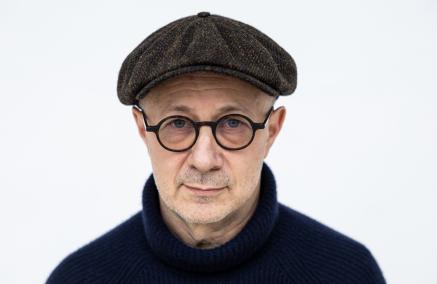 Mikołaj Grynberg - mężczyzna ubrany na czarno, na nosie ma okulary w ciemnych oprawkach, a na głowie szary kaszkiet.