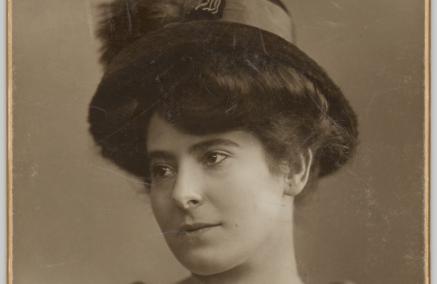 Fotografia portretowa Heleny Rubinstein w półprofilu. Kobieta jest ubrana w bluzkę z delikatnym wzorem, z dekoltem w kształcie litery V, wykończonym niewielkim bukietem żywych rumianków. Głowę przystraja kapelusz z wąskim, zaokrąglonym w dół rondem, z szeroką wstążką.