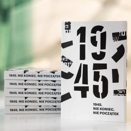 Na stole stoi katalog wystawy "1945. Nie koniec nie początek", obok na stosie leży kilka egzemplarzy publikacji. W tle ściana w holu głównym Muzeum POLIN.