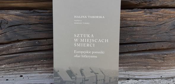 Okładka książki Halina Taborska (współpraca Marian Turski) "Sztuka w miejscach śmierci. Europejskie pomniki ofiar hitleryzmu". Książka oparta o ścianę ze starych drewnianych belek