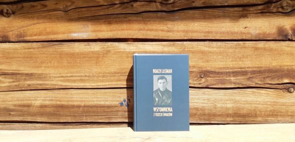 Okładka książki Noah Lasman "Wspomnienia z trzech światów". Książka oparta o ścianę starego drewnianego domu