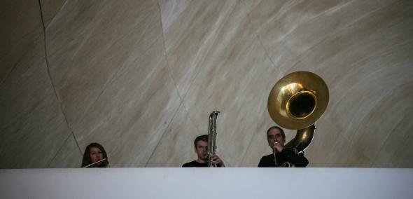 Na zdjęciu widać trzy osoby, trzymające instrumenty dęte, n.in. puzon i tubę, wyglądające zza parapetu na balkonie wewnętrznym w Muzeum POLIN