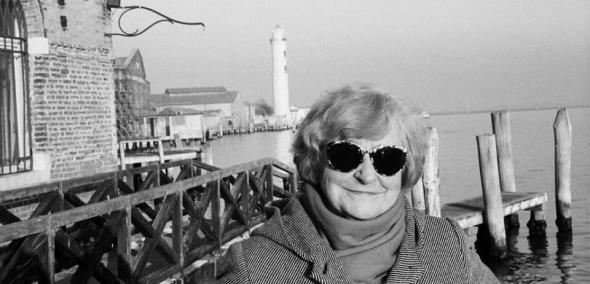 Uśmiechnięta kobieta na tle drewnianego pomostu na morzu. Z lewej strony fragment ceglanego budynku. W oddali latarnia morska.
