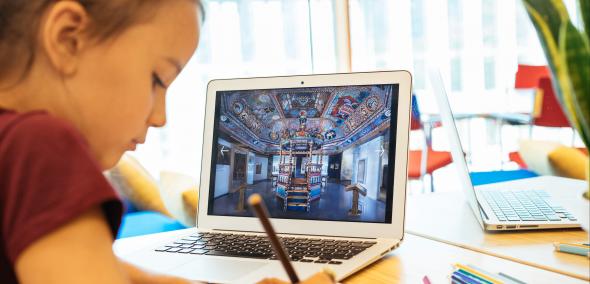Dziecko - z lewej strony ekranu - siedzi przez otwartym laptopem i notuje w zeszycie